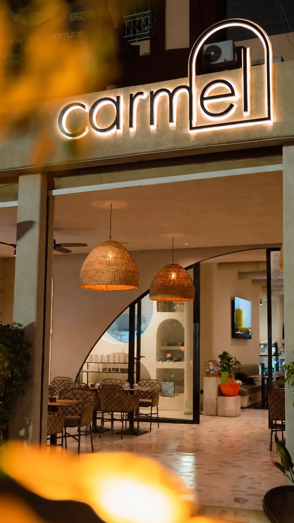 The entrance of Carmel Marrakech, its illuminated sign and the dining room under wicker pendant lights
